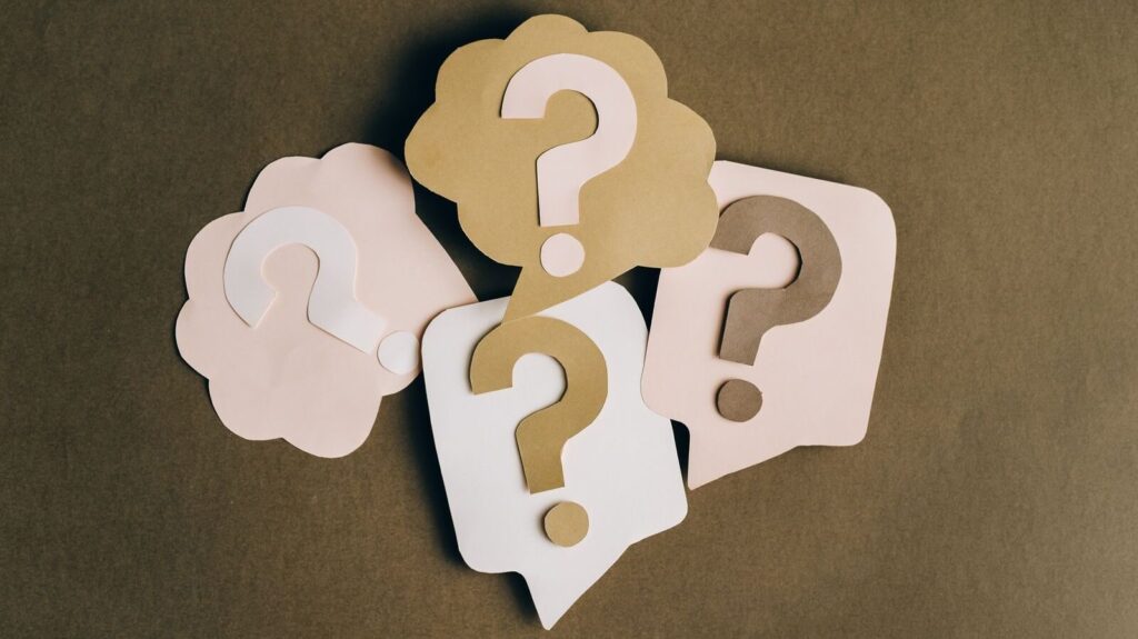 Four paper cutouts of question marks in beige and white on a brown surface, symbolizing inquiry and curiosity.