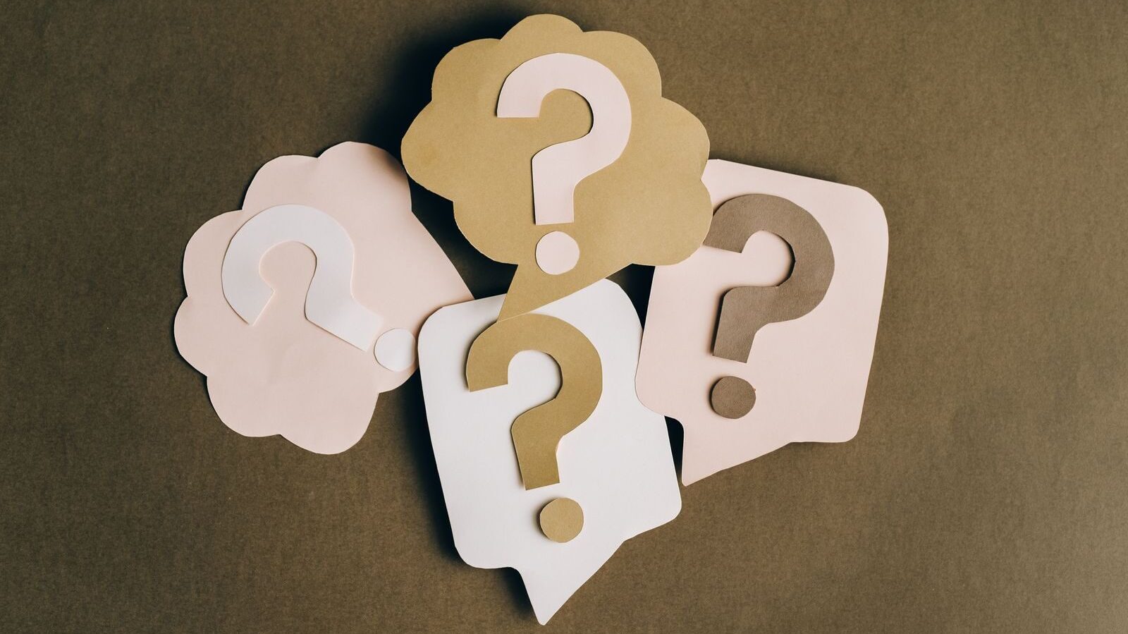Four paper cutouts of question marks in beige and white on a brown surface, symbolizing inquiry and curiosity.